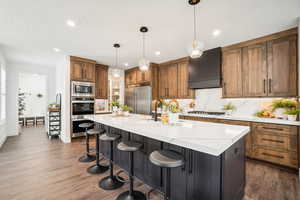 Kitchen with premium range hood, a kitchen island with sink, dark wood-type flooring, built in appliances, and pendant lighting