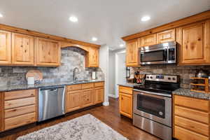 Kitchen with stainless steel appliances, backsplash, dark stone countertops, sink, and dark hardwood / wood-style flooring