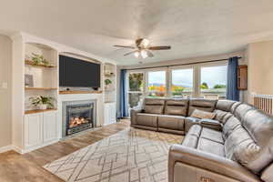 Living room featuring light wood-type flooring, a fireplace, a textured ceiling, ceiling fan, and built in shelves