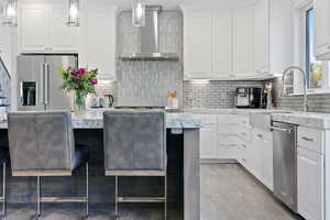 Kitchen with appliances with stainless steel finishes, white cabinetry, hanging light fixtures, and wall chimney range hood