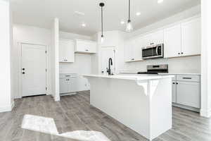 Kitchen with light wood-type flooring, a center island with sink, hanging light fixtures, appliances with stainless steel finishes, and white cabinets