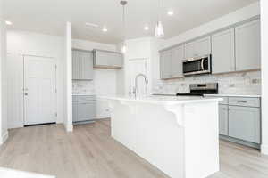 Kitchen featuring light hardwood / wood-style flooring, gray cabinets, stainless steel appliances, hanging light fixtures, and a kitchen island with sink