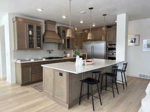 Kitchen featuring a large island, backsplash, stainless steel appliances, custom exhaust hood, and light wood-type flooring