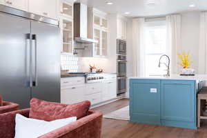 Kitchen with dark wood-type flooring, wall chimney exhaust hood, white cabinets, built in appliances, and decorative backsplash