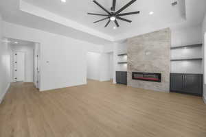 Unfurnished living room featuring ceiling fan, a raised ceiling, light wood-type flooring, a fireplace, and built in shelves