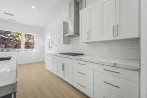 Kitchen with wall chimney range hood, light stone countertops, stainless steel gas cooktop, white cabinetry, and light hardwood / wood-style floors