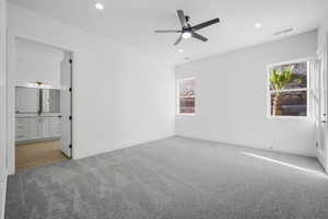 Unfurnished bedroom featuring multiple windows, ensuite bath, light colored carpet, and ceiling fan