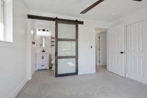 Unfurnished bedroom featuring connected bathroom, ceiling fan, light colored carpet, and a barn door