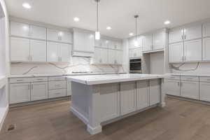 Kitchen featuring oven, light stone counters, an island with sink, dark wood-type flooring, and decorative light fixtures