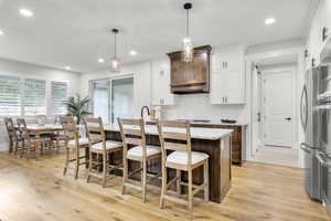 Kitchen with light wood-type flooring, backsplash, white cabinetry, pendant lighting, and a kitchen island with sink