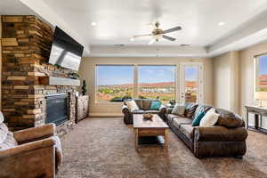 Carpeted living room featuring a stone fireplace, a healthy amount of sunlight, a raised ceiling, and ceiling fan