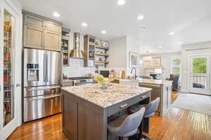 Kitchen featuring wood-type flooring, wall chimney exhaust hood, a kitchen island, light stone countertops, and appliances with stainless steel finishes