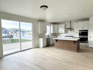 Kitchen featuring light wood-type flooring, wall chimney exhaust hood, stainless steel appliances, a center island, and white cabinetry