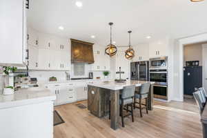 Kitchen featuring stainless steel appliances, white cabinetry, light LVP, a kitchen island, and premium range hood, double oven.