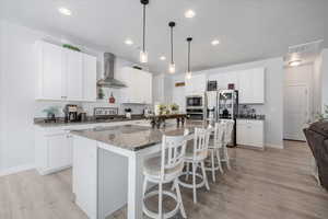 Kitchen with white cabinets, wall chimney exhaust hood, light hardwood / wood-style flooring, an island with sink, and appliances with stainless steel finishes