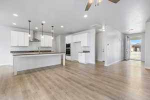 Kitchen featuring a kitchen island with sink, wall chimney range hood, dark countertops, and white cabinetry