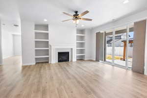 Unfurnished living room featuring built in shelves, light wood-style flooring, a tiled fireplace, ceiling fan, and baseboards