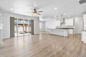 Kitchen with wall chimney exhaust hood, open floor plan, a kitchen island with sink, white cabinetry, and pendant lighting