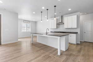 Kitchen with visible vents, white cabinetry, hanging light fixtures, wall chimney range hood, and a center island with sink