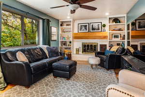 Living room with a brick fireplace, built in shelves, wood-type flooring, and ceiling fan