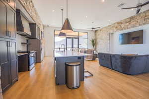 Kitchen with light wood-type flooring, decorative light fixtures, stove, wall chimney exhaust hood, and a kitchen island with sink