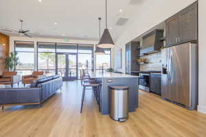 Kitchen with stainless steel appliances, light hardwood / wood-style floors, hanging light fixtures, a kitchen island with sink, and wall chimney range hood