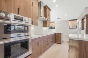 Kitchen featuring stainless steel double oven, wall chimney range hood, tasteful backsplash, and black electric cooktop