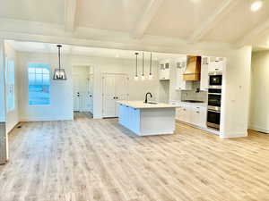 Kitchen featuring premium range hood, sink, beam ceiling, white cabinetry, and an island with sink