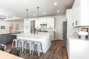 Kitchen with appliances with stainless steel finishes, dark hardwood / wood-style floors, hanging light fixtures, white cabinets, and a kitchen island with sink