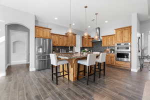 Kitchen featuring a center island with sink, extractor fan, appliances with stainless steel finishes, decorative light fixtures, and dark hardwood / wood-style flooring