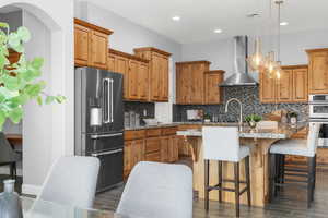 Kitchen featuring dark wood-type flooring, a kitchen island with sink, wall chimney range hood, pendant lighting, and appliances with stainless steel finishes