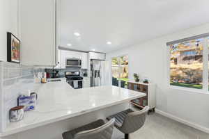 Kitchen featuring kitchen peninsula, light stone counters, appliances with stainless steel finishes, a kitchen bar, and white cabinets