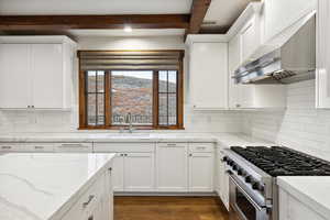 Kitchen with wall chimney range hood, stainless steel stove, sink, white cabinets, and beamed ceiling