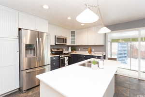 Kitchen featuring white cabinetry, stainless steel appliances, decorative light fixtures, and an island with sink