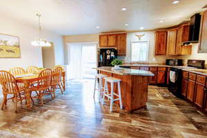 Kitchen with a breakfast bar, black appliances, a center island, wall chimney range hood, and pendant lighting