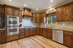 Chef's Dream  Kitchen featuring stainless steel appliances, decorative backsplash, sink, custom exhaust hood, and light hardwood flooring