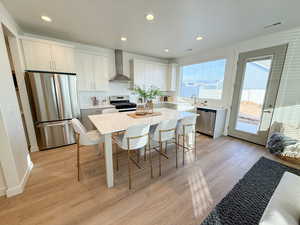 Kitchen featuring a kitchen breakfast bar, wall chimney exhaust hood, stainless steel appliances, a center island, and light hardwood / wood-style floors