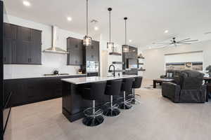 Kitchen with light wood-type flooring, wall chimney exhaust hood, stainless steel appliances, a center island with sink, and hanging light fixtures