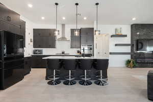 Kitchen with double oven, black fridge, a kitchen island with sink, and wall chimney range hood