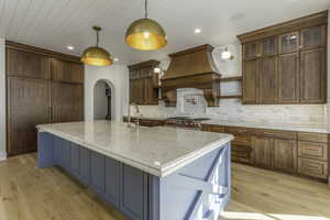 Kitchen with premium range hood, light stone counters, decorative light fixtures, light hardwood / wood-style floors, and an island with sink