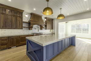 Kitchen with sink, premium range hood, pendant lighting, light hardwood / wood-style floors, and a center island with sink