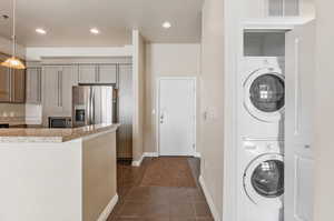 Kitchen featuring stacked washer and clothes dryer, light stone counters, stainless steel appliances, dark tile patterned floors, and decorative light fixtures