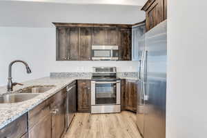 Kitchen with dark brown cabinetry, sink, appliances with stainless steel finishes, light stone countertops, and light hardwood / wood-style flooring