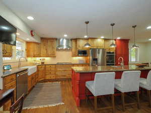 Kitchen with a center island with sink, appliances with stainless steel finishes, hanging light fixtures, dark wood-type flooring, and wall chimney range hood