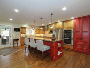 Kitchen with appliances with stainless steel finishes, wall chimney exhaust hood, decorative light fixtures, a kitchen island with sink, and dark wood-type flooring