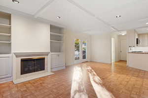 Unfurnished living room featuring built in shelves, a premium fireplace, coffered ceiling, crown molding, and light parquet flooring