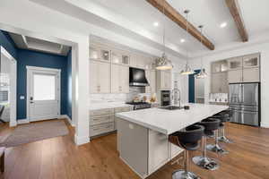 Kitchen featuring ventilation hood, a center island with sink, appliances with stainless steel finishes, pendant lighting, and beamed ceiling