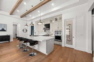 Kitchen featuring light hardwood / wood-style floors, beamed ceiling, an island with sink, pendant lighting, and exhaust hood