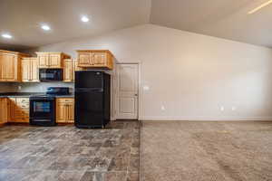 Kitchen with light brown cabinetry, black appliances, lofted ceiling, and dark carpet