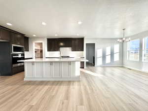 Kitchen featuring stainless steel appliances, light hardwood / wood-style floors, a notable chandelier, an island with sink, and dark brown cabinets